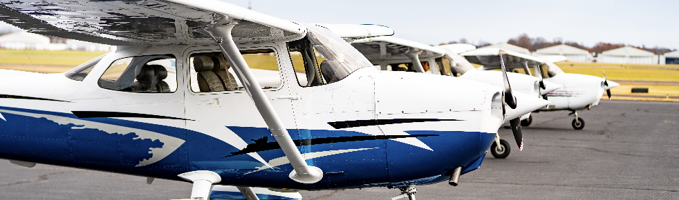 Row of small white and blue propeller airplanes parked on an airstrip, nose to the right, with hangars in the background.